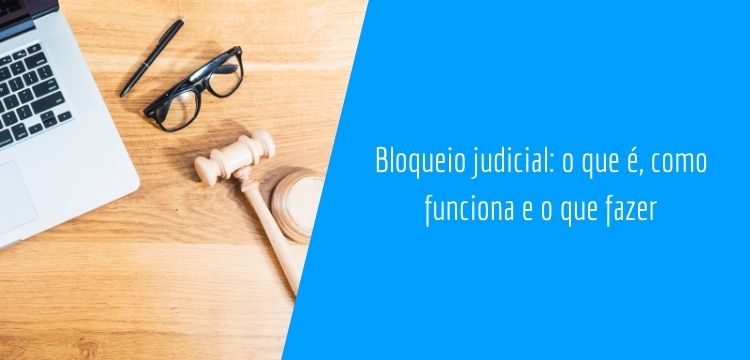 imagem retangular. na parte direita encontramos um polígono azul turquesa com o título do texto "bloqueio judicial: o que é, como funciona e o que fazer", na metade esquerda da imagem, temos um martelo judiciário, um óculos retangular de aro preto, uma caneta preta e um notebook, todos com vista superior em uma mesa de madeira.