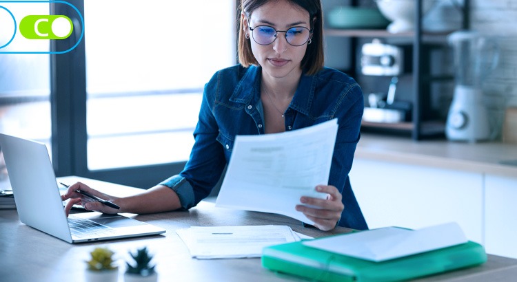 Uma mulher vestida com camisa jeans lendo um relatório na frente do notebook.