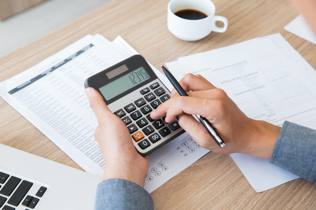 a mesa com uma calculadora sendo segurada por duas mãos que realizam uma conta, como se estivesse calculando o COFINS, há papéis espalhados e no canto esquerdo há uma parte de um notebook e no canto direito há uma xícara de café