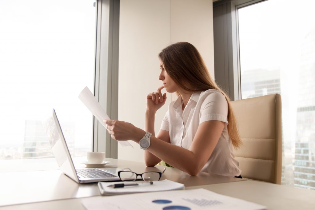 mulher branca sentada em uma mesa segurando um papel e colocando a mão no rosto com tom de dúvida, na mesa que ela está sentada há um laptop, papéis, xícara de café, óculos e canela, a foto está em tons de bege 