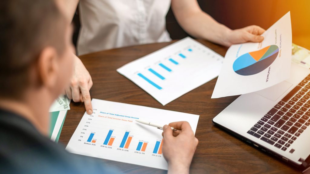 mesa de madeira com duas pessoas sentadas, em cima da mesa há folhas de gráficos e planejamento, notebook, as pessoas estão realizando um orçamento empresarial 