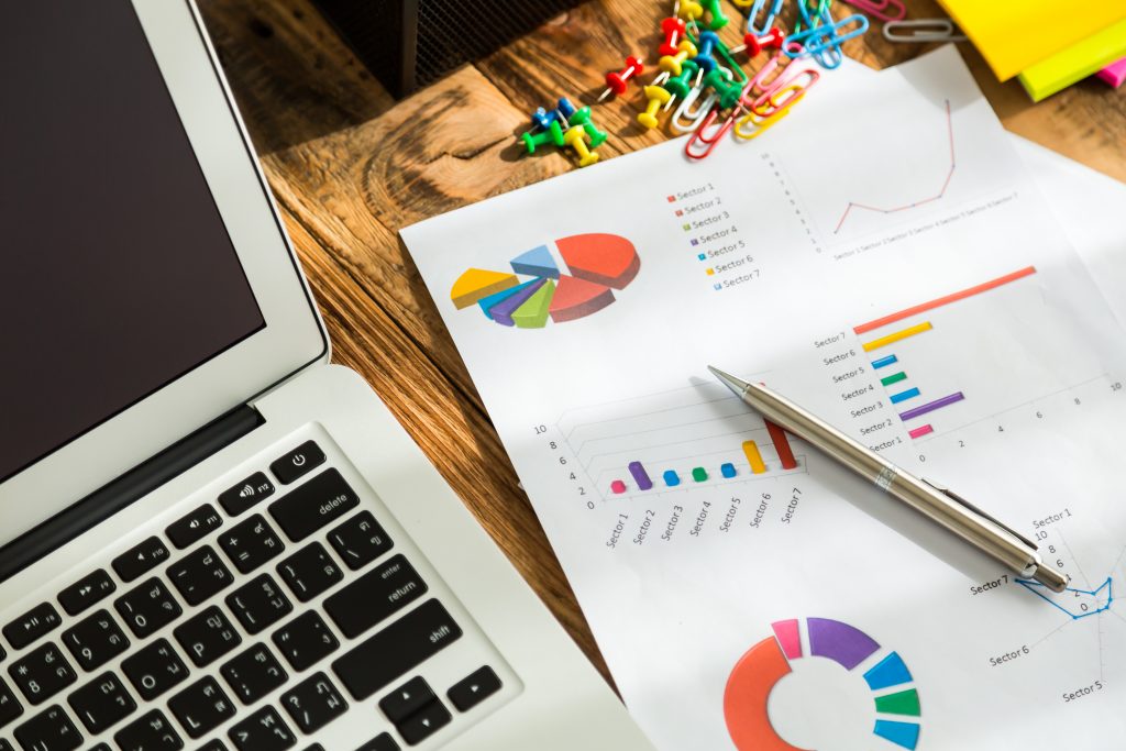 computador em cima de uma mesa de madeira com um papel com gráficos coloridos e uma caneta demonstrando KPIs