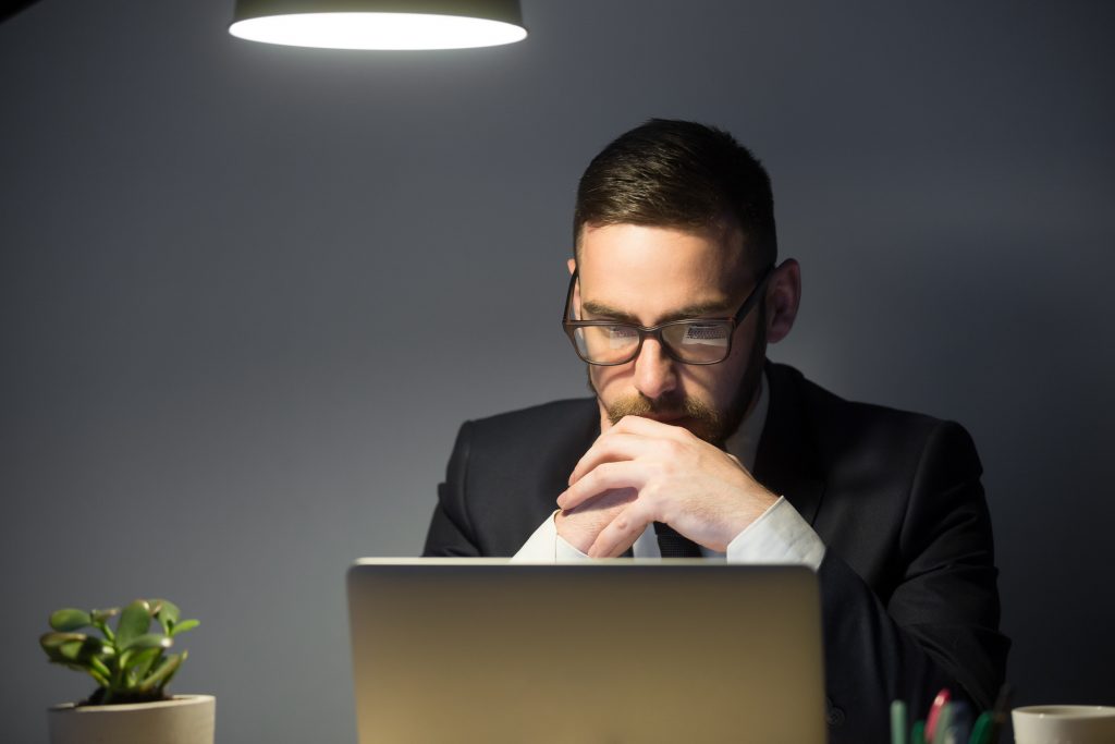 homem branco de terno sentado em frente á um computador, ao fundo há um vaso com uma planta verde e ao teto há uma luminária