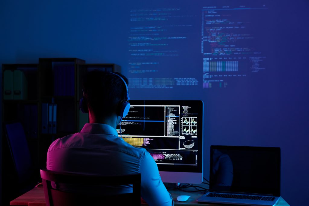 homem usando fones de ouvido está sentando de frente para um monitor com vários abas abertas e com fundos pretos, a parede ao fundo tem uma projeção dos códigos e do lado direito há um notebook