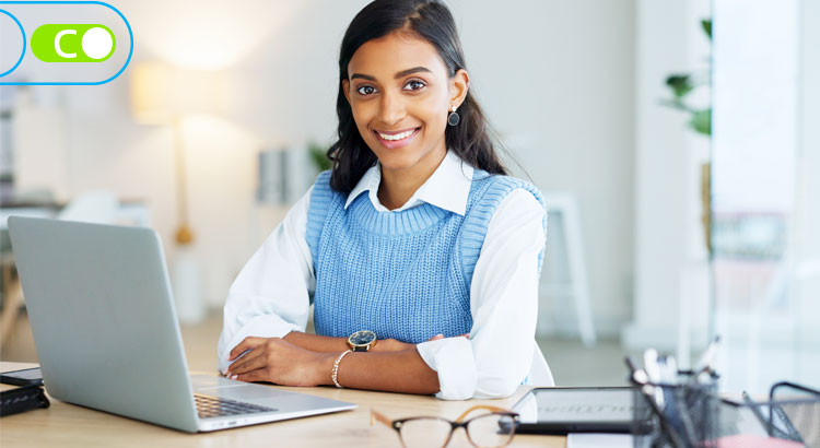 Na foto, uma mulher jovem, vestida com camisa branca e um colete azul, com os braços cruzados em cima da mesa, a sua frente na mesa, há um notebook, um óculos e alguns papéis.