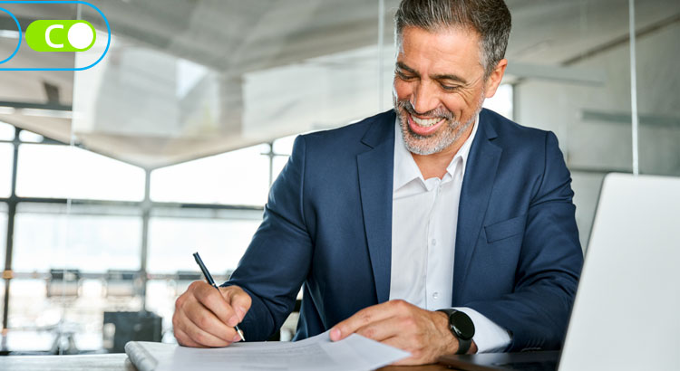 Na imagem, um homem branco e com barba grisalha, vestindo terno azul e camisa branca, está sentado a frente de uma mesa, assinando papéis e sorrindo.