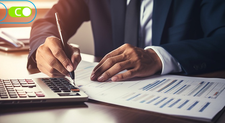 Na imagem há um homem de terno assinando um papel em cima de uma mesa e existe uma calculadora ao lado dele.