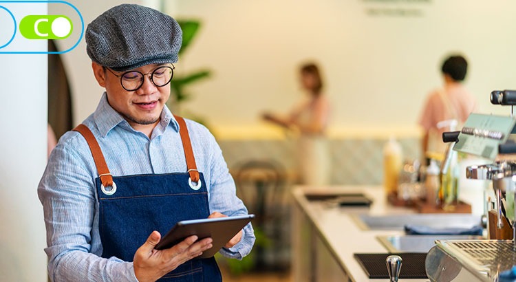 Na imagem, há um homem vestido com camisa azul e um avental jeans, ele também está usando boina e óculos. Está olhando para um tablet o qual está segurando com as mãos e ao fundo o que parece ser uma bancada de um café.