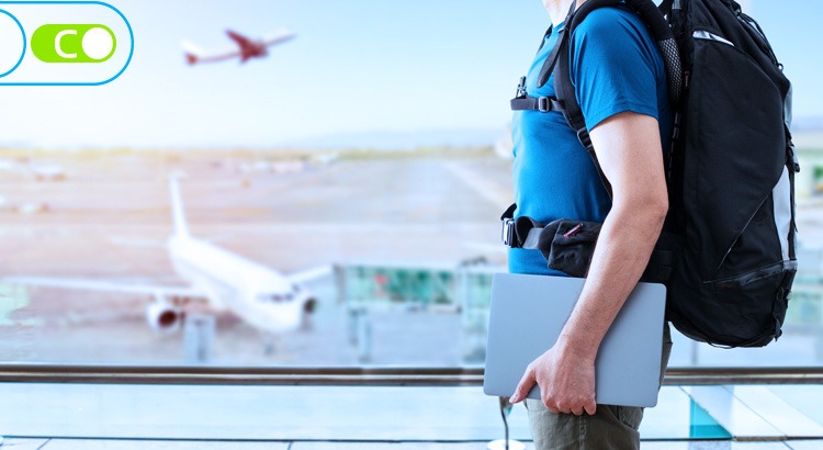 Você está vendo uma foto de um homem com camiseta azul, de lado, segurando um notebook em sua mão, junto ao corpo e e com uma mochila de viagem. Ao fundo é uma pista de aeroporto, com aviões prontos para decolar e um avião decolando.
