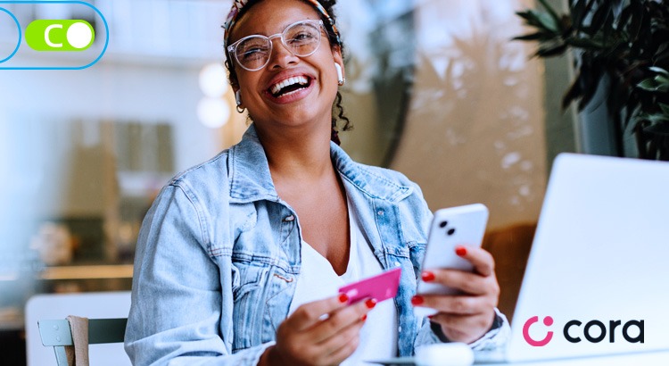 Na imagem, uma mulher negra, com os cabelos presos por uma bandana, sorrindo, ela usa óculos e está com uma jaqueta jeans, segurando em sua mão direita um cartão da Cora e em sua mão esquerda um celular.