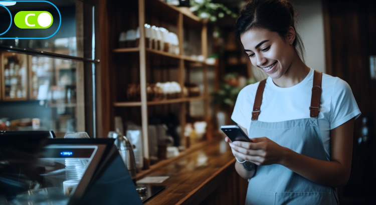 Na imagem, uma mulher, vestida de avental e touca, atrás de um balcão de uma loja. Ela está segurando um celular e olhando para ele com um sorriso no rosto.