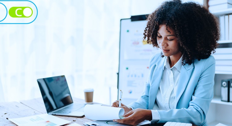 Na foto, uma mulher negra com um lindo cabelo black power escrevendo em um papel na mesa, na frente dela a um notebook e ela está de roupa social.